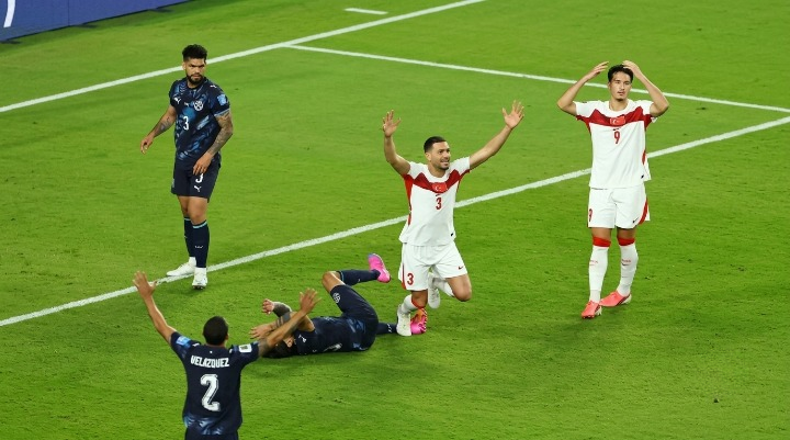 Turkish football players in white kits celebrating and reacting on a green pitch with a crowded stadium background.